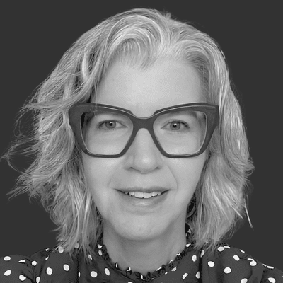 A black and white photograph of a Melissa Reeve. Melissa is wearing large framed glasses and a spotty blouse. She is smiling at the camera.