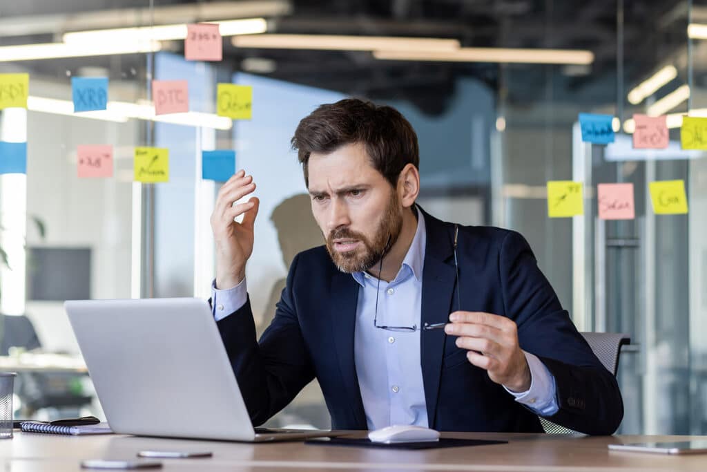A businessman in a suit looks stressed while working on his laptop in a modern office. Sticky notes with various business terms are visible on the glass wall behind him, highlighting a busy work